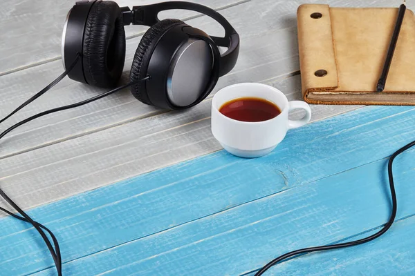 Black headphones with cord, brown notebook in leather cover, pencil and cup of tea on blue and white wooden tabletop. Close up, copy space — Stock Photo, Image