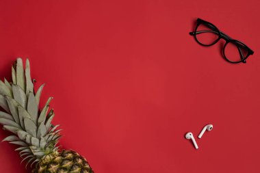 Siyah çerçeveli şık gözlükler, kablosuz kulaklıklar veya kulaklıklar ve kırmızı stüdyo arka planında olgun ananas. Kapat, boşluğu kopyala