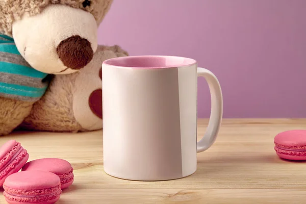 Bílý porcelánový hrnek na kávu nebo čaj na dřevěném stolku vedle plyšového medvídka a macaronů na růžovém pozadí studia. Zavřít, kopírovat mezeru — Stock fotografie
