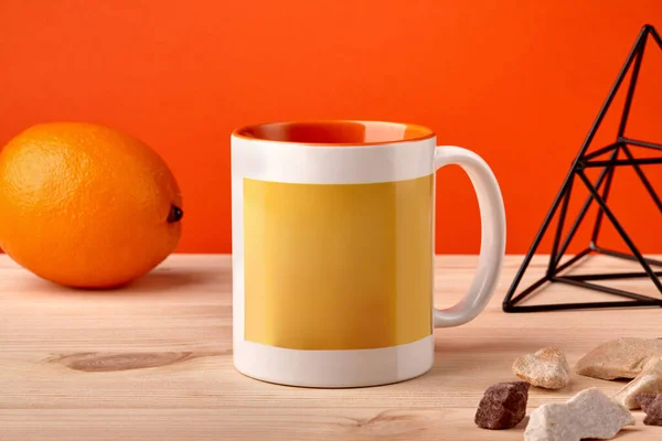 Barevný porcelánový hrnek na dřevěné desce vedle oranžové, kovová figurka trojúhelníku a malé kameny na oranžovém pozadí. Zavřít, kopírovat mezeru — Stock fotografie