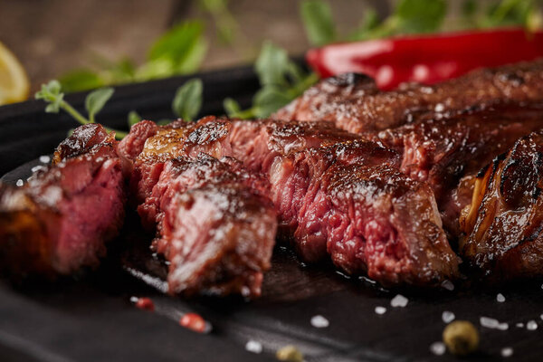 Closeup of slices of spicy juicy beef striploin steak on black wooden platter with blurred background of fresh greens and red hot pepper