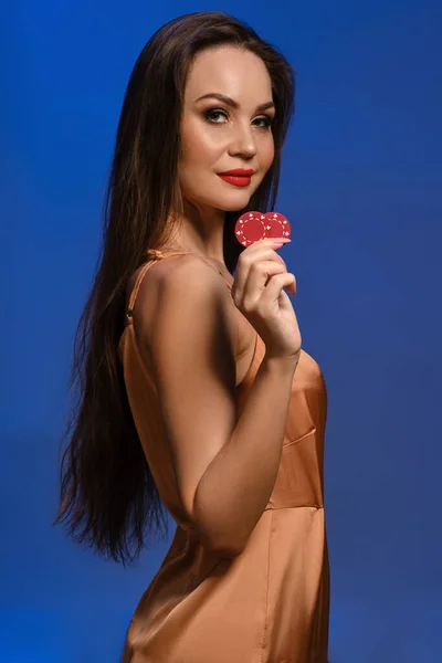 Young brunette female in silk beige dress. She smiling, showing two red chips, posing sideways on blue background. Poker, casino. Close-up — Stock Photo, Image