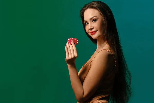 Bruneta modelka v hedvábných béžových šatech. Usmíval se, ukazoval dva červené hranolky, pózoval bokem na zeleném pozadí studia. Poker, kasino. Detailní záběr — Stock fotografie