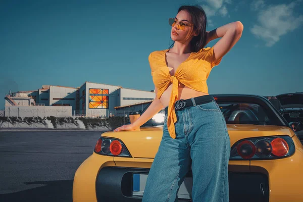 Mulher de jeans azul, top laranja e óculos de sol está posando perto de cabriolet carro amarelo com xícara de papel de café no tronco no estacionamento. Espaço de cópia — Fotografia de Stock