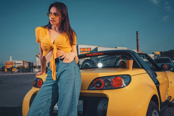 Lady em jeans azul, top laranja e óculos de sol está posando inclinado no roadster carro amarelo com xícara de papel de café no porta-malas no estacionamento. Fechar — Fotografia de Stock