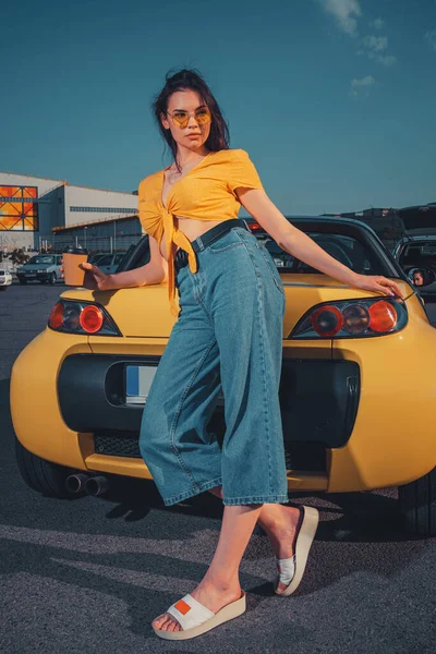 Modelo jovem em jeans azul, top laranja e óculos de sol. Ela está segurando xícara de papel de café, apoiando-se no carro amarelo no estacionamento. Comprimento total, mock up — Fotografia de Stock