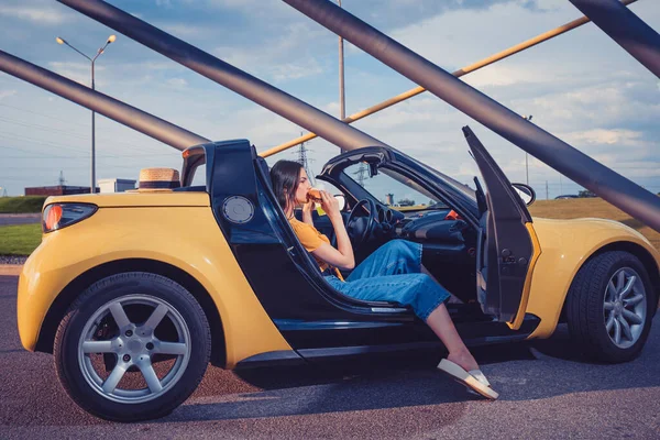 Junges Mädchen in blauen Jeans, orangefarbenem Oberteil und Flip Flops isst Hamburger, während sie im gelben Auto-Cabrio sitzt. Fast Food, ungesunde Ernährung — Stockfoto