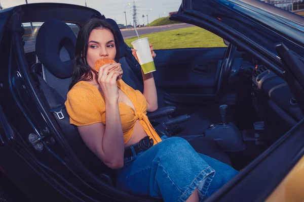 Modelo em jeans azuis e top laranja está comendo hambúrguer e segurando bebida em copo de papel enquanto sentado em cabriolet carro amarelo. Comida rápida. Espaço de cópia — Fotografia de Stock
