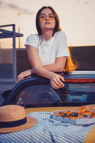 Jovem senhora em óculos de sol, t-shirt branca está posando em cabriolet carro amarelo com pizza, pano listrado e chapéu em seu tronco. Comida rápida. Fechar — Fotografia de Stock