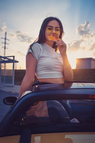 Jovem fêmea de óculos de sol e camiseta branca está comendo pizza enquanto posando em pé no cabriolet carro amarelo. Comida rápida. Pôr-do-sol. Fechar — Fotografia de Stock