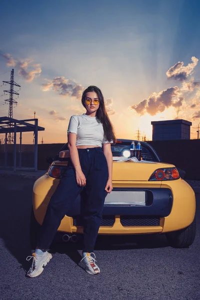 Feminino em óculos de sol, top branco, jeans azul e tênis está posando ao ar livre perto do carro amarelo com duas garrafas de vidro de refrigerante em seu tronco. Faz asneira. — Fotografia de Stock
