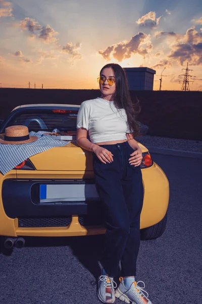 Молодая леди в солнечных очках, в белом топе, синих джинсах и кроссовках позирует на улице возле желтого авто-родстера с пиццей на багажнике. Фаст-фуд — стоковое фото
