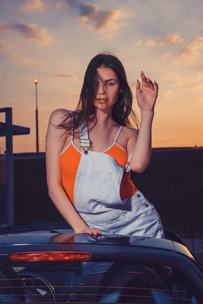 Sexy jovem fêmea em branco geral e laranja t-shirt está posando ao ar livre em pé no carro cabriolet. Noite de verão, pôr-do-sol. Fechar — Fotografia de Stock