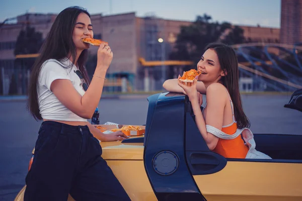 De jolies jeunes femmes en tenue décontractée mangent de la pizza tout en posant en cabrio jaune avec des frites et de l'eau gazeuse sur son tronc. Restauration rapide — Photo