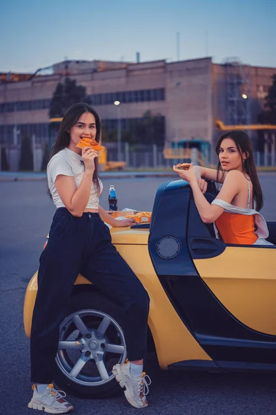 Lindas fêmeas em roupas casuais comendo pizza, posando em carro amarelo com batatas fritas e refrigerante em garrafa de vidro em seu tronco. Comida rápida. Comprimento total — Fotografia de Stock
