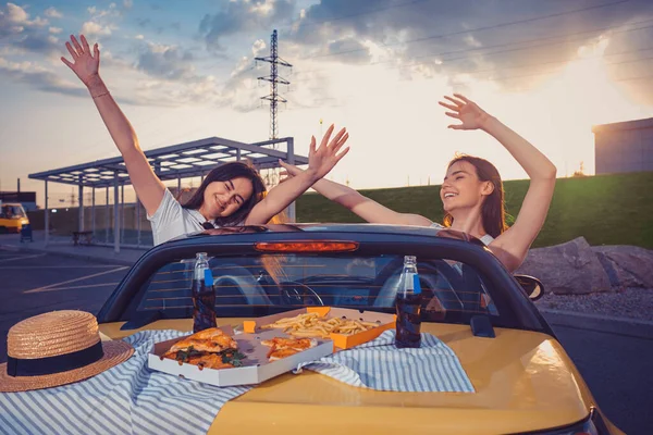 Junge Mädchen lächeln, heben die Hände und posieren im gelben Auto mit Pommes, Pizza und Limo in Glasflaschen auf dem Kofferraum. Fast food. Kopierraum — Stockfoto