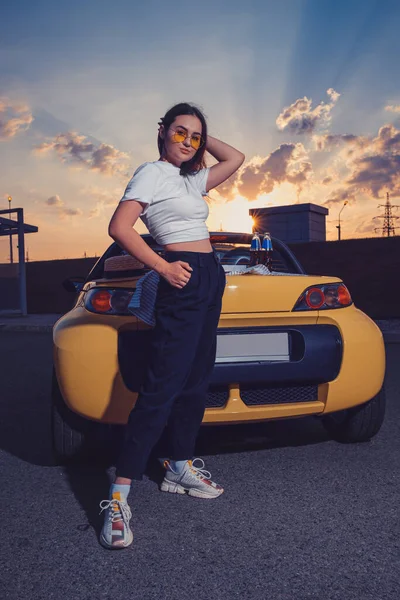 Chica atractiva en gafas de sol, top blanco, pantalones negros y zapatillas de deporte está posando al aire libre cerca de cabriolet coche amarillo con dos botellas de refresco en su maletero —  Fotos de Stock