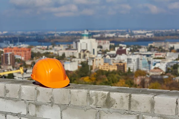 Casque de sécurité en plastique orange est sur un mur de briques. Toit du bâtiment moderne en construction. Paysage urbain panoramique flou, ciel bleu, journée ensoleillée — Photo