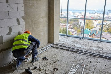 İş giysisi ve güvenlik kaskı yapımcısı, panoramik pencereli inşaat halindeki binanın dairesine plastik borular kuruyor. Yüksek katlı ev.