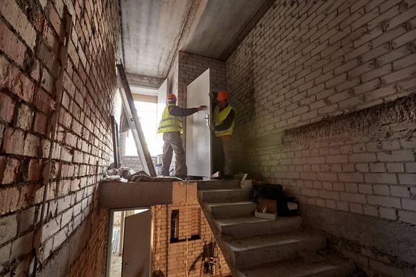 Two workers in workwear and hard hats are installing a door in an unfinished stairwell of a high-rise building under construction