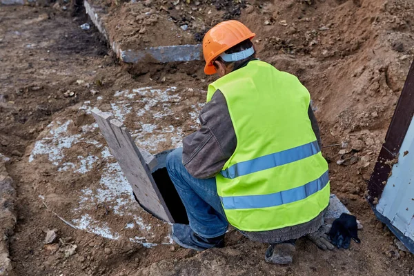 Le travailleur est à la recherche dans une trappe d'égout pour vérifier les communications menant à un nouveau bâtiment de grande hauteur en construction. Gros plan, vue arrière — Photo