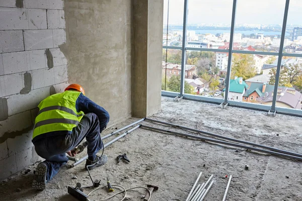 Constructeur en vêtements de travail et casque de sécurité est d'installer des tuyaux en plastique dans l'appartement de l'immeuble en construction avec des fenêtres panoramiques. Maison de grande hauteur — Photo