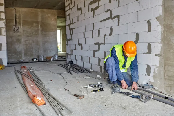 Constructeur en vêtements de travail et casque de sécurité orange installe des tuyaux en plastique à l'aide d'outils modernes dans un appartement du bâtiment en construction — Photo