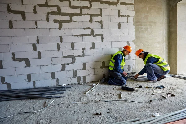 Deux constructeurs masculins en vêtements de travail et chapeaux durs de sécurité installent des tuyaux en plastique à l'aide d'outils modernes dans un appartement de bâtiment en construction — Photo