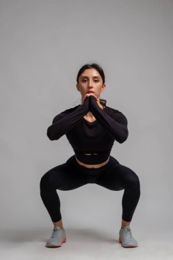 Atletik esmer, gri arka planda vücut ağırlığı çömelmeleri yapıyor.