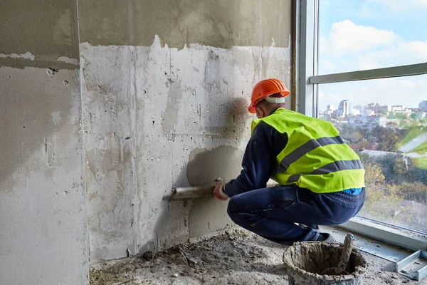 Travailleur qualifié en vêtements de travail et chapeau dur orange met du mastic sur le mur, en utilisant la spatule. Appartement d'un immeuble en construction — Photo