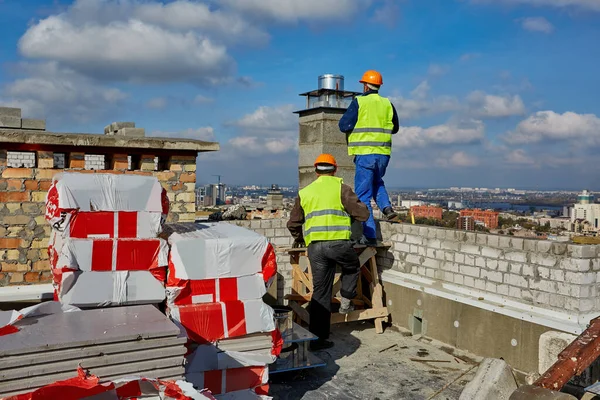 Deux hommes constructeurs professionnels en vêtements de travail et hardhats orange travaillent avec un système de ventilation sur le toit du bâtiment en construction — Photo