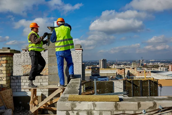 Deux travailleurs professionnels masculins portant des vêtements de protection et des casques de sécurité travaillent avec un système de ventilation sur le toit du bâtiment en construction — Photo