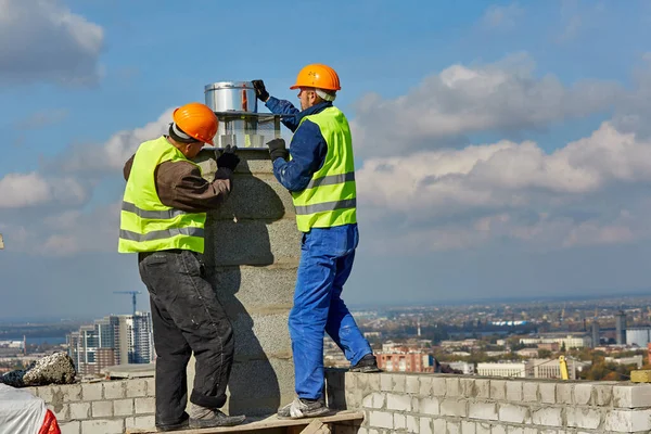 Deux travailleurs en vêtements de protection et des casques de sécurité installent un tuyau pour le système de ventilation sur le toit du bâtiment moderne en construction — Photo