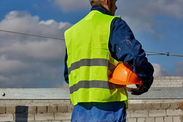 Constructeur professionnel en vêtements de travail tient un casque orange tout en se tenant sur le toit du bâtiment en construction et en regardant loin. Ciel bleu — Photo