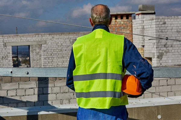 Mature worker in protective clothing is holding an orange safety helmet while standing on the roof of building under construction. Blue sky