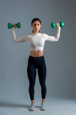 Genç sporcu kadın gri arka planda dambıl gibi omuzlara baskı yapıyor.