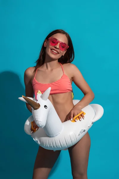 Chica sonriente en traje de baño y gafas de sol sobre fondo azul con anillo de goma unicornio — Foto de Stock