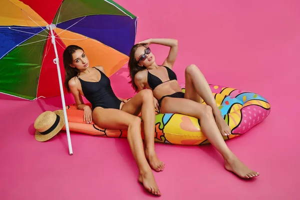 Mädchen in Badeanzügen sitzen auf Luftmatratzen unter Sonnenschirm auf rosa Hintergrund — Stockfoto