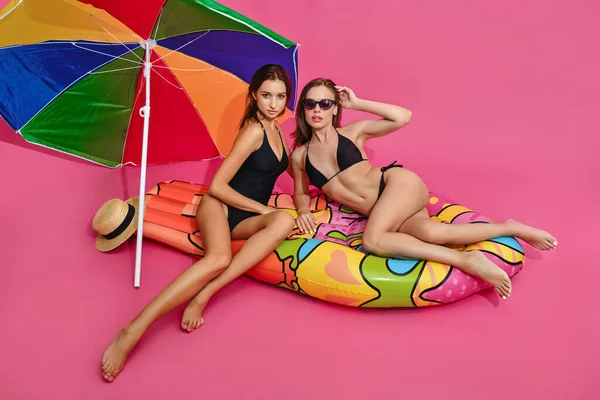 Frauen in Badeanzügen posieren auf Luftmatratze neben Sonnenschirm auf rosa Hintergrund — Stockfoto
