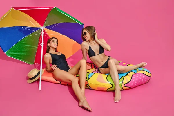 Fröhliche Mädchen in Badeanzügen auf aufblasbarer Matratze unter Sonnenschirm auf rosa Studiohintergrund — Stockfoto