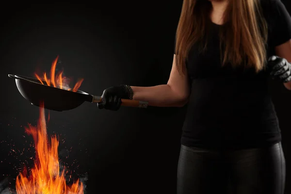 Morena senhora com as mãos tatuadas, vestida com leggings e t-shirt. Segurando uma panela de wok acima do fogo contra o fundo preto. Conceito de cozinha. Vista lateral — Fotografia de Stock