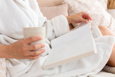 Beyaz bornozlu genç kadın, yatak odasında kahve içip dergi ya da kitap okuyor. Yakın plan.