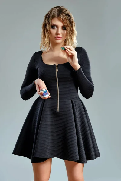 Mujer rubia en vestido corto negro que muestra algunas fichas de colores, posando sobre fondo gris. Entretenimiento, póquer, casino. Primer plano. — Foto de Stock