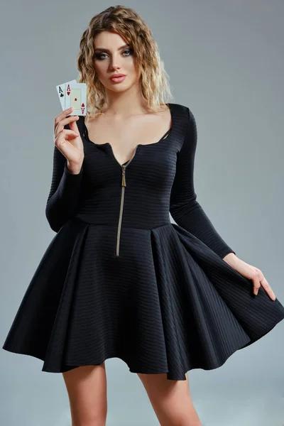 Mujer rubia en vestido corto negro mostrando dos cartas de juego, posando sobre fondo gris. Entretenimiento, póquer, casino. Primer plano. — Foto de Stock