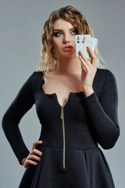 黒のドレスのブロンドの女性は、灰色の背景に対してポーズを2枚のトランプを示しています。ギャンブルエンターテイメント,ポーカー,カジノ.閉鎖. — ストック写真