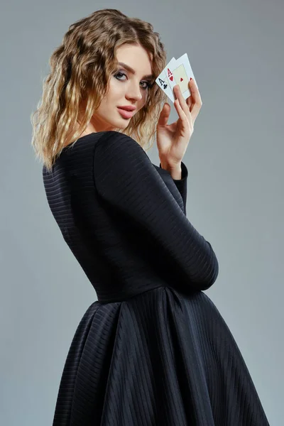 Blond žena v černých šatech ukazuje dvě hrací karty, pózovat bokem na šedém pozadí. Hazardní hry, poker, kasino. Detailní záběr. — Stock fotografie