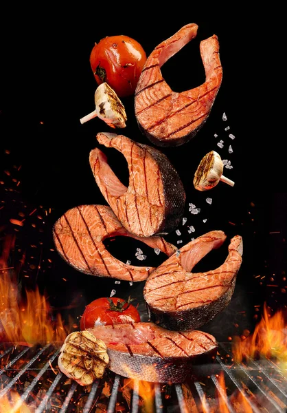 Des steaks de saumon grillés aux tomates, à l'ail et au sel tombent sur fond noir. Barbecue barbecue grill, feu, charbon et fumée. Gros plan — Photo