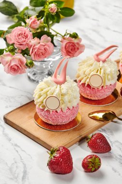 Beyaz krema, çikolata ve taze çilekle süslenmiş pembe kekler, pembe gül vazosunun yanındaki tahta tahtada oturuyorlar.