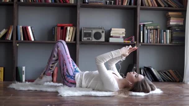 Arka planda bir şömine ile evde yerde yatarken bir smartphone üzerinden müzik dinleme kadın — Stok video