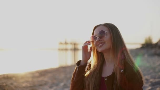 Joven hermosa chica en gafas de sol en la puesta del sol sonriendo en la cámara — Vídeos de Stock